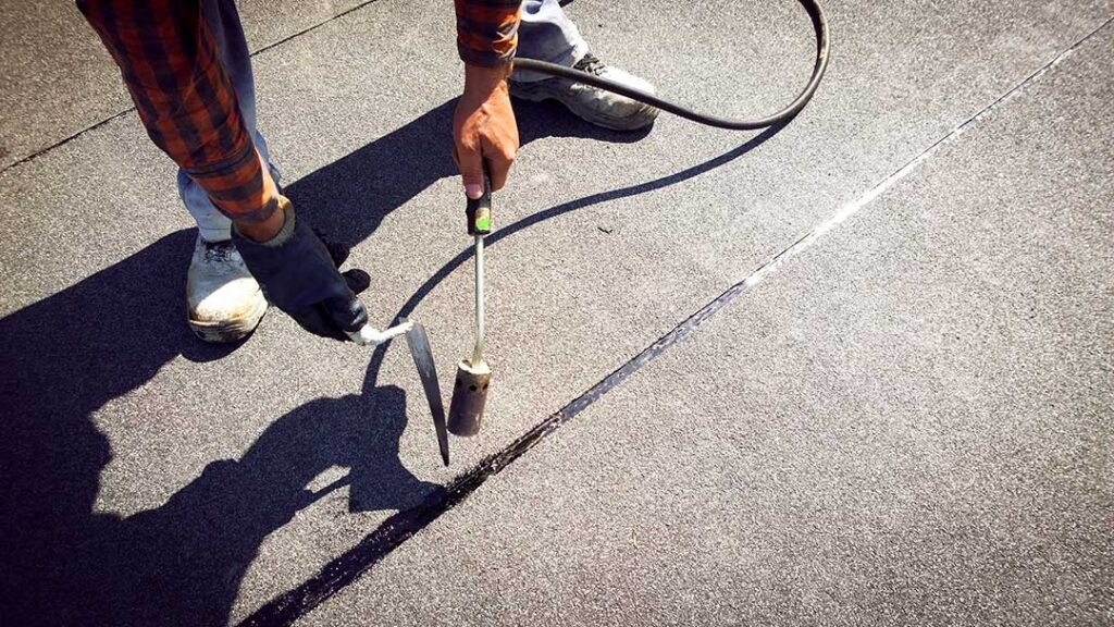 Person using a soft roof rake to clear leaves from a low-slope shingle roof.